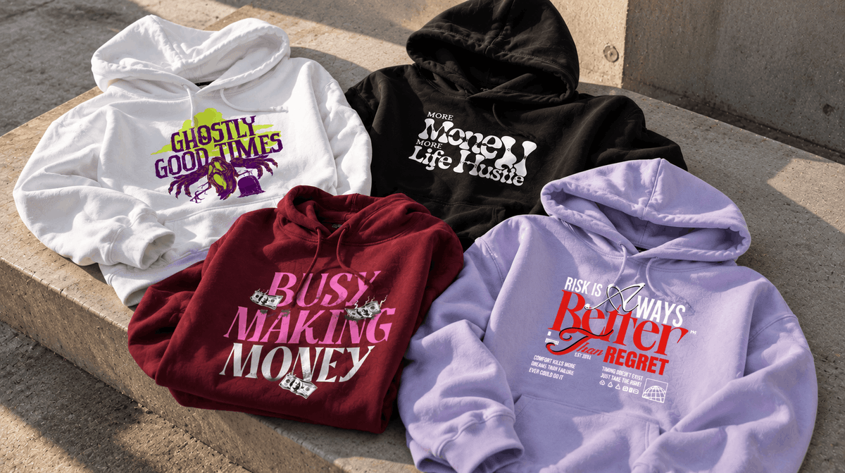 Four hoodies in different colors are arranged on concrete outdoors. The hoodies are white, black, burgundy, and light purple, each with a stylized graphic text print on the front.